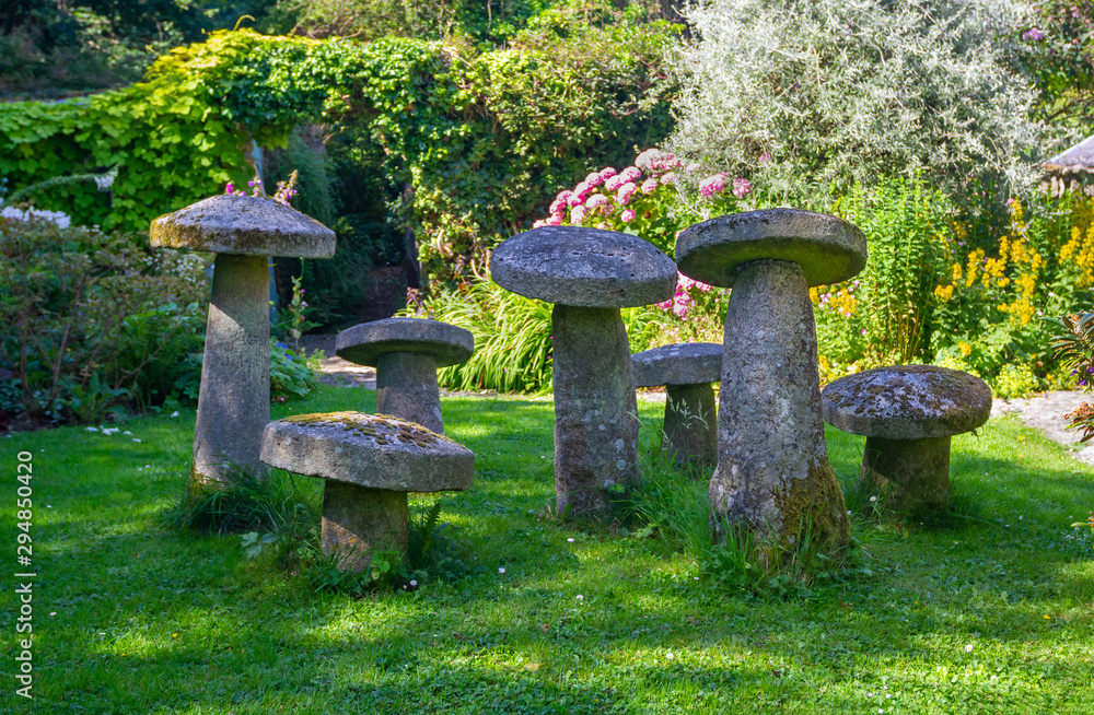 Seven stone staddle stones, ornamental toadstools, in sun lit summer garden. Old, weathered fairy stones covered with lichen and moss. Also called mushroom stones. Garden in Ireland. Seven stone staddle stones, ornamental toadstools, in sun lit summer garden. Old, weathered fairy stones covered with lichen and moss. Also called mushroom stones. Garden in Ireland.
