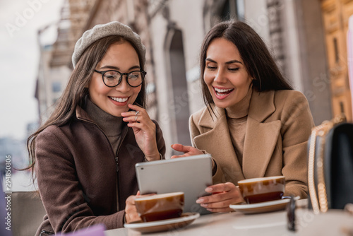 Portrait of smiling two young women at a cafe table looking in digital tablet