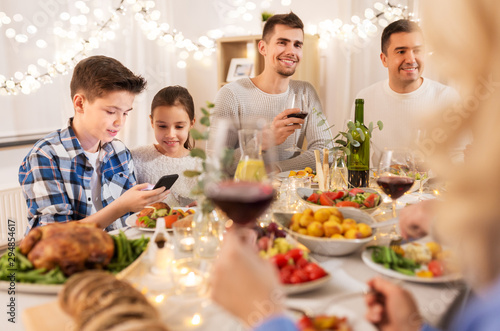 technology, holidays and people concept - happy children with smartphone at family dinner party