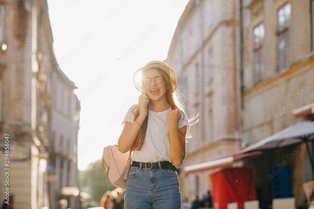 Fashionable hipster girl in stylish hat, talks with boyfriend via cell phone, shares happy emotions after exploring new sights in city, stands over building outdoor