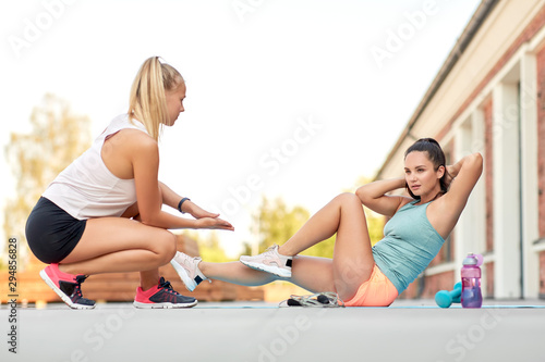 fitness, sport and healthy lifestyle concept - young woman assisting her friend doing bicycle crunches on mat outdoors