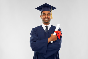 education, graduation and people concept - happy smiling indian male graduate student in mortar board and bachelor gown with diploma over grey background