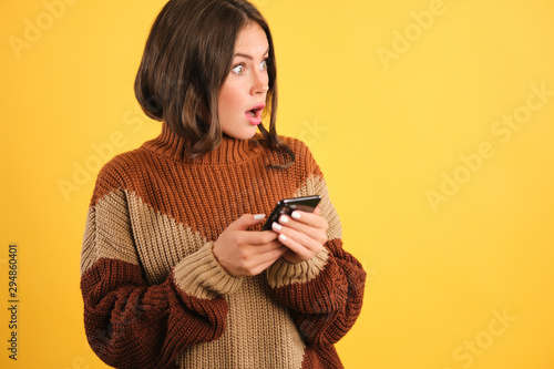 Surprised girl in sweater with cellphone amazedly looking aside over yellow background photo