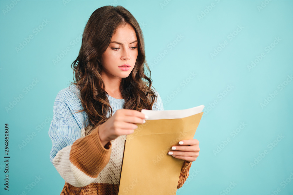 Pensive girl in sweater thoughtfully opening response envelope over blue background Pensive girl in sweater thoughtfully opening response envelope over blue background