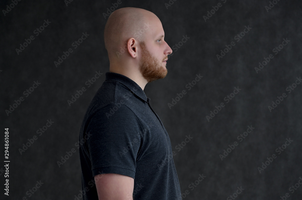 Side portrait of a young bald man in a gray T-shirt on a gray background. It s right without emotion.