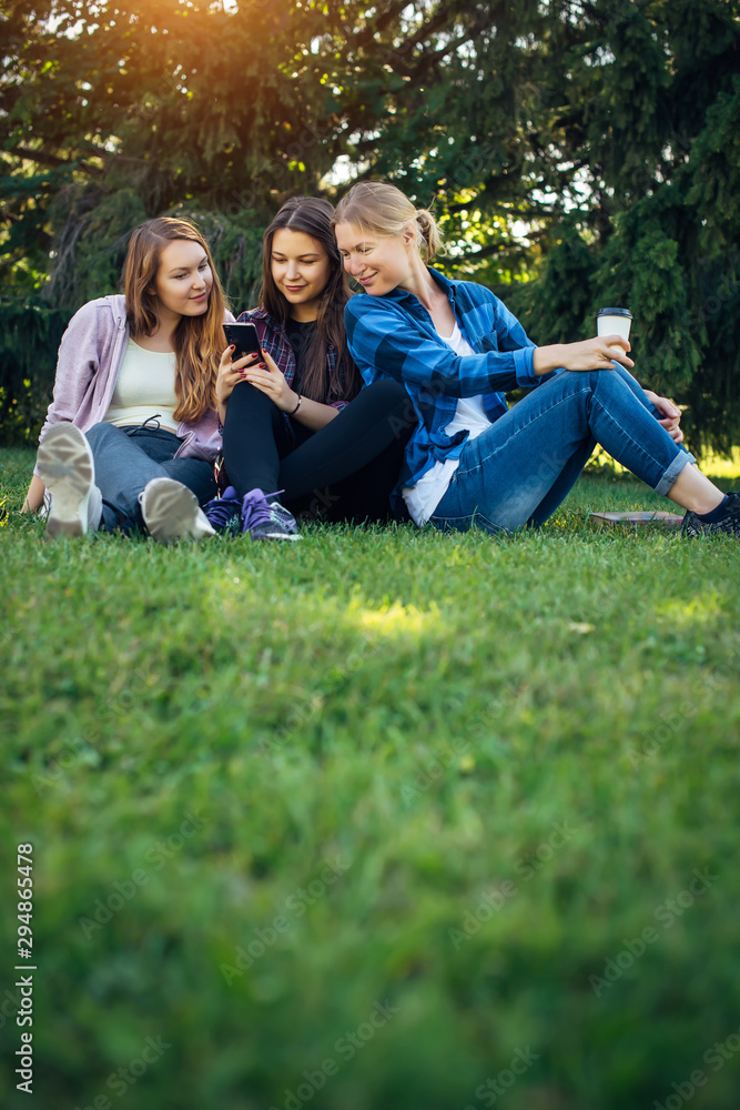 Three friends spend their free time sitting in the park on green lawn. Young pretty girls talking, smiling, looking photo at smartphone. Concept of female friendship. Vertical image. Three friends spend their free time sitting in the park on green lawn. Young pretty girls talking, smiling, looking photo at smartphone. Concept of female friendship. Vertical image.