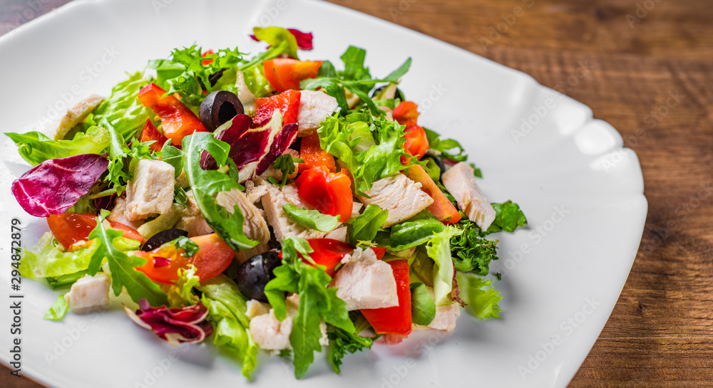 Fresh salad with chicken breast, arugula, black olives,red pepper, lettuce, fresh sald leaves and tomato on a white plate on wooden table background Fresh salad with chicken breast, arugula, black olives,red pepper, lettuce, fresh sald leaves and tomato on a white plate on wooden table background