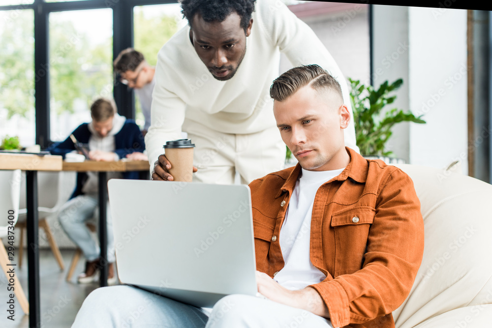 african american businessman with coffee to go looking at laptop in hands of colleague