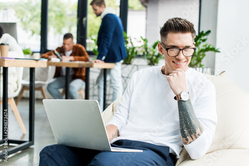 young businessman in glasses using laptop and smiling at camera near colleagues working in office
