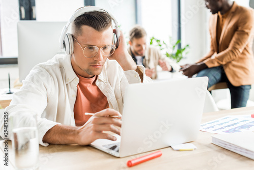 young businessman in headphones using laptop while working near multicultural colleagues