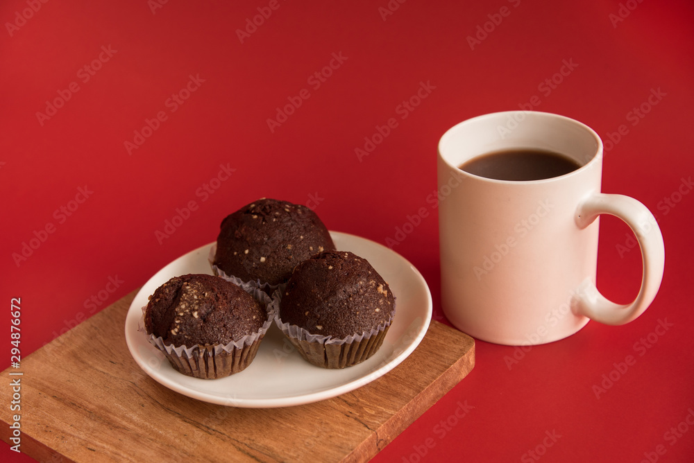 A white Cup of coffee stands on a wooden Board. Chocolate cupcakes are on the plate. Red background.