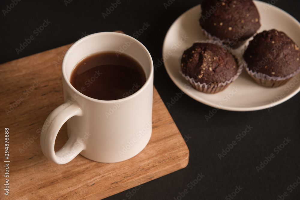 A white Cup of coffee stands on a wooden Board. Chocolate cupcakes are on the plate. Black background. A white Cup of coffee stands on a wooden Board. Chocolate cupcakes are on the plate. Black background.