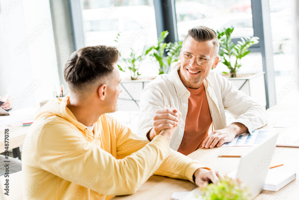 Fototapeta premium two young businessmen shaking hands while sitting together at workplace