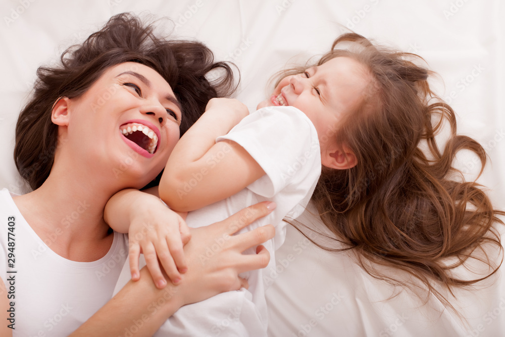 Portrait of brunette long hair mother and daughter on a white linen bed. The concept of a happy family life, relaxation, comfort, fun. View from above... Portrait of brunette long hair mother and daughter on a white linen bed. The concept of a happy family life, relaxation, comfort, fun. View from above...