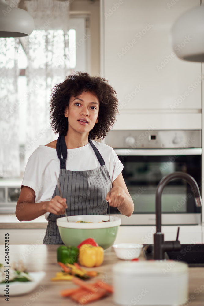Attractive mixed race woman in apron and with curly hair mixing vegetables in bowl while standing in kitchen at home. Attractive mixed race woman in apron and with curly hair mixing vegetables in bowl while standing in kitchen at home.