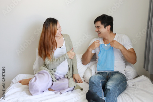medium shot asian pregnant woman and caucasian husband holding baby shirt with love happy smiling face on bed in bedroom