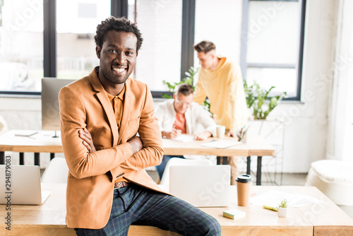 young african american businessman smiling at camera while sitting on desk in office