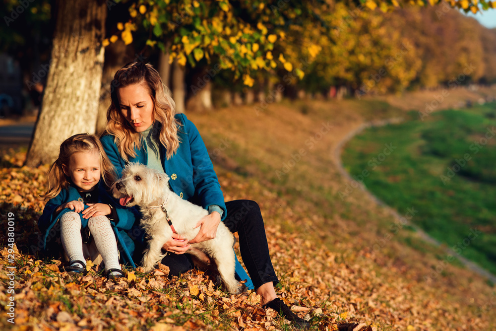 Fototapeta premium Happy mom with her child relaxing with white terrier dog on autumn leaves. Autumn season time. Fashion, family, autumn, lifestyle