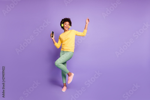 Full length body size photo of trendy stylish cute free girlfriend wearing green pants trousers yellow sweater footwear in headphones listening to music dancing isolated violet pastel color background
