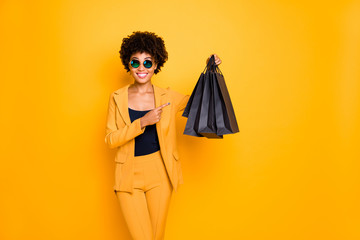 Look real bargain. Portrait of positive brunette curly hair afro american girl hold black bags point index finger show what she shopped bought wear style pants outfit isolated yellow color background