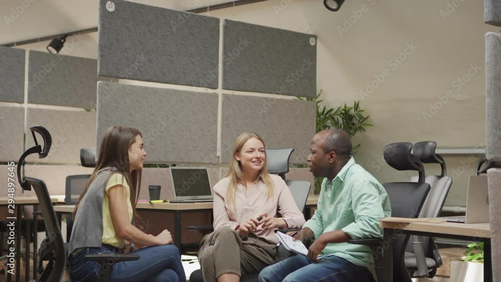Bald African American male with notebook sitting on chair and discussing project with female coworkers in cubicle of modern office Bald African American male with notebook sitting on chair and discussing project with female coworkers in cubicle of modern office