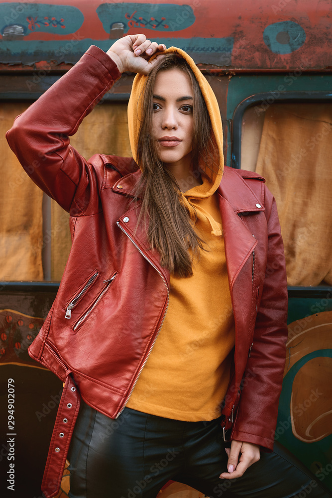 Stylish woman in colored leather biker jacket and hoodie posing in the hood with colored bus on background.