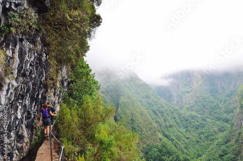 Hikers walking on a narrow path on the edge of the rock during Levada do Caldeirao Verde Trail. Misty green mountains in background. Dangerous hiking. Portuguese tourist attraction. Fog, foggy