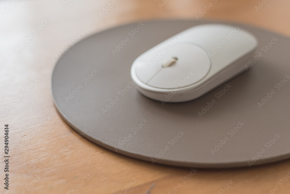 White and silver wireless mouse on a gray round leather mouse pad on a wooden surface. White and silver wireless mouse on a gray round leather mouse pad on a wooden surface.