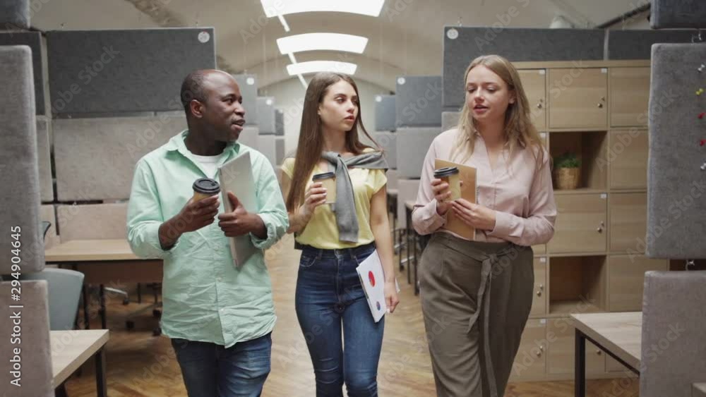 Tracking shot of diverse man and women with takeaway cups of hot beverages walking and talking with each other while working in modern office together Tracking shot of diverse man and women with takeaway cups of hot beverages walking and talking with each other while working in modern office together