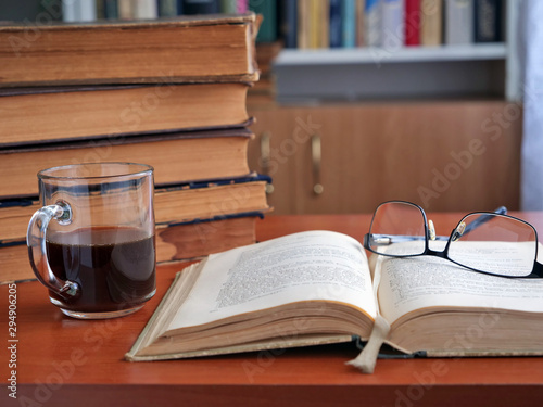 Open old book and glasses on the table with a cup of coffee. Bookcase in the background in a living room.