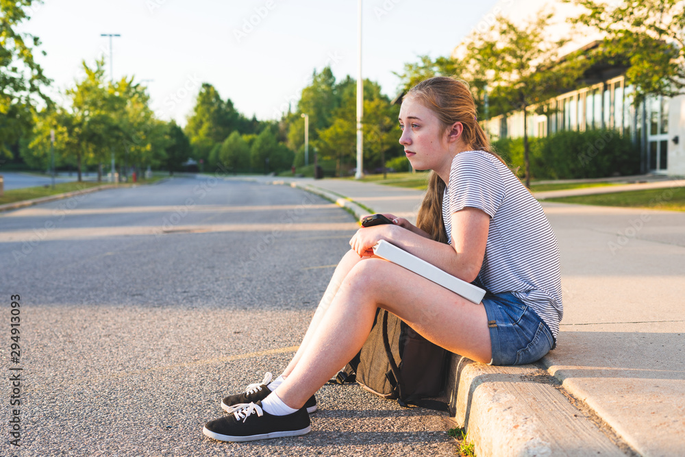 Depressed/Sad teen girl sitting on a curb in front of a high school during sunset while sitting next to a backpack, holding binders, and looking at a smartphone.