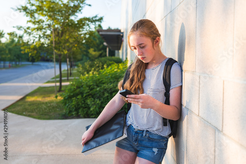 Depressed/Sad teen girl leaning against high school wall during sunset while wearing a backpack and holding binders/smartphone.