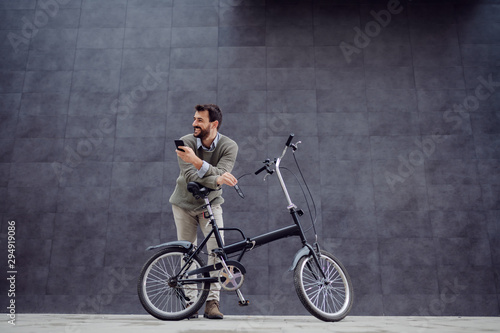 Full length of smiling handsome caucasian fashionable man leaning on his bicycle and holding smart phone and glasses. In background is gray wall.