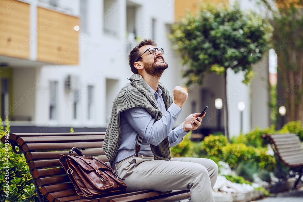 Fototapeta premium Successful handsome fashionable caucasian businessman sitting on bench in park, holding smart phone and rejoicing for achievement. Next to him is leather handbag.