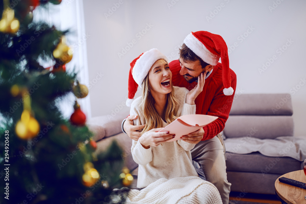 Obraz premium Amazed cute Caucasian blonde woman sitting on sofa in living room, hugging her boyfriend and receiving gift. Both having santa hats on heads. In foreground is christmas tree. Living room interior.