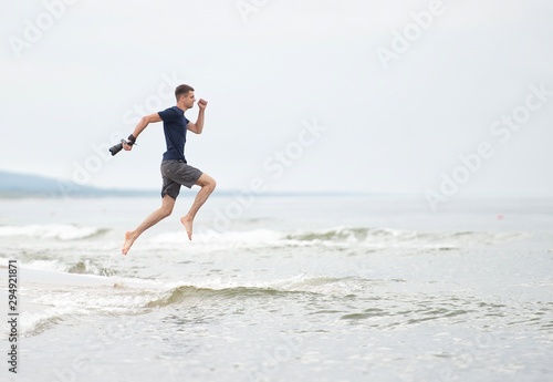 happy young guy in shorts and a t-shirt with a camera in his hand, running through the waves, graceful shot in a jump, in hot weather on the shore of the resort, bright bright frame