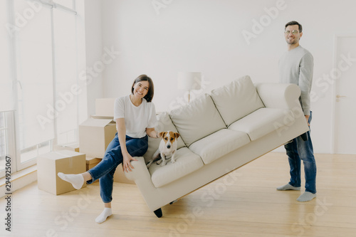 фотографія Happy couple move furniture in their new modern home, carry sofa with pet, pose