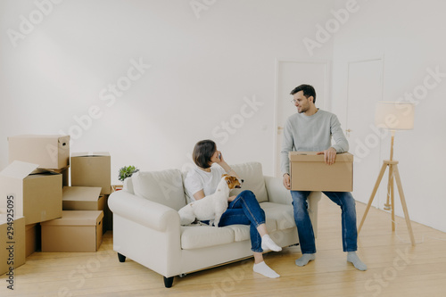 Positive woman and man pose in empty spacious room during relocation day, husband carries cardboard boxes with belongings, wife has telephone conversation, sits on comfortable sofa with dog. © VK Studio
