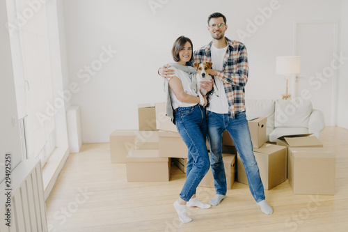 Young couple in love stand closely to each other, embrace and holds favourite dog, pose in empty room, surrounded with cardboard boxes, make family portrait in new apartment. Moving Day concept
