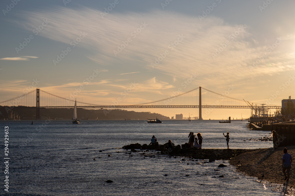 People taking pictures near the river Tagus in Lisbon with the 25th of April Bridge in the background.