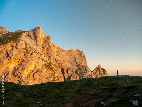 Hiker standing on the edge of grassy hill in the Alps raises her arms in victory