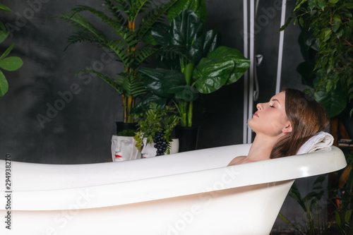 A young woman lies with her eyes closed in a white graceful bathtub, where green plants are nearby. Take a bath in the tropical area