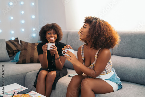 Two Young african american black women relaxing drinking cup of hot coffee or tea on couch at home.