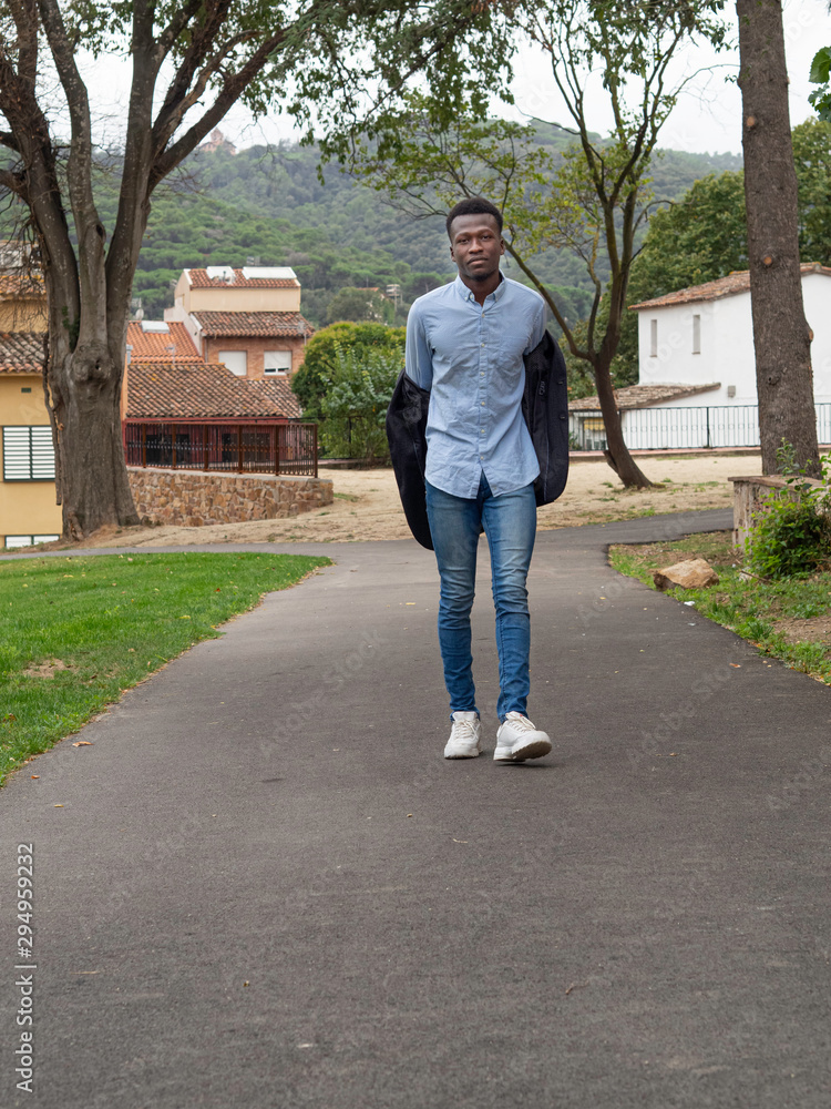 Hombre joven de Senegal quitándose la chaqueta mientras camina con camisa clara arrugada , pantalones vaqueros estrechos y zapatillas foto de | Adobe Stock