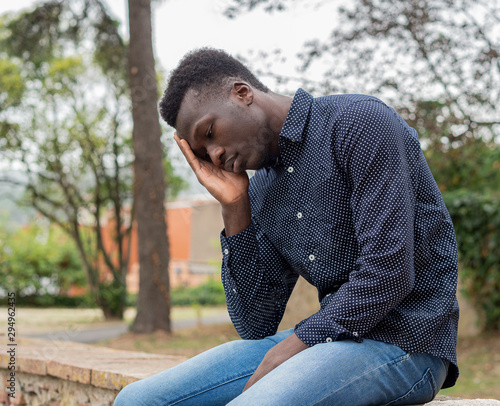 Hombre joven negro de Senegal con pantalones vaqueros y camisa azul, sentado, con una mano en la cabeza y triste photo