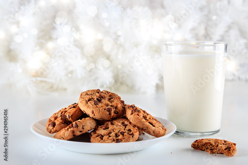 Closeup to delicious handmade chocolate chip cookies on a white plate and a glass of fresh milk on Christmas day. The lights are on decorating the white Christmas tree. Christmas holidays concept