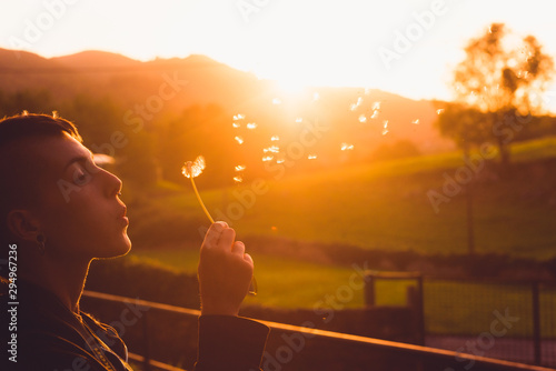 A young and attractive girl with short hair remains pierced while blowing a dandelion at sunset.