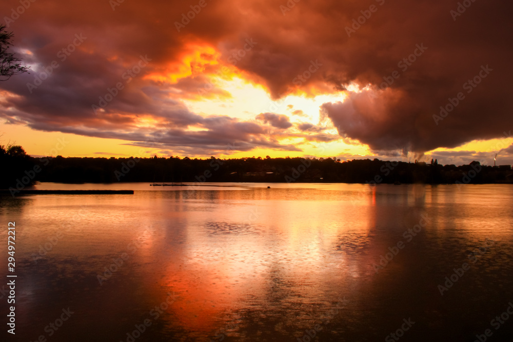Dramatic sky at sunset over water with a pier in the foreground in the middle of a lake. Silhouette of forest in the background. 