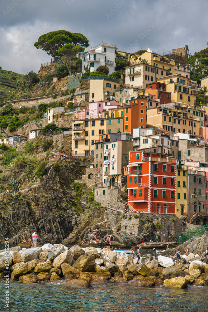 Riomaggiore, Cinque Terre, Italy - August 17, 2019: Beautiful colorful houses on the rocky coast of the Ligurian Sea. Spreading tree on the mountain. Tourists sunbathe on the rocks
