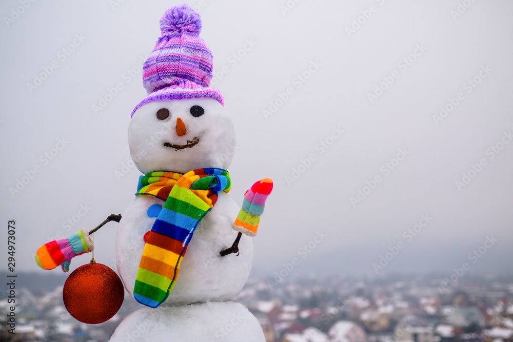 Obraz premium Snowman holds new year ball. Snow man in winter hat. Christmas background with snowman. Funny snowman in stylish hat and scarf. Smiling snow man with Christmas toy. Merry Christmas and Happy new year.
